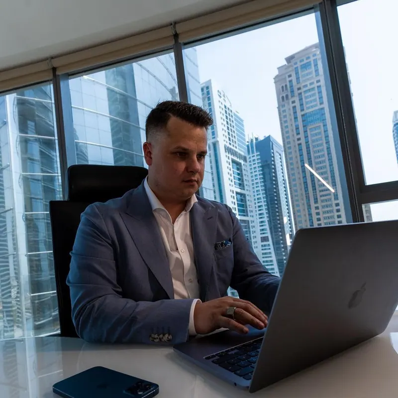 Dennis Loos im Anzug am Laptop in seinem Büro vor der Skyline von Dubai