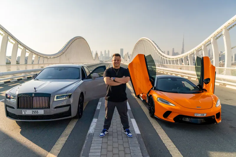 Dennis Loos mit verschränkten Armen zwischen einem silbernen Rolls-Royce und einem orangefarbenen McLaren auf einer Brücke in Dubai